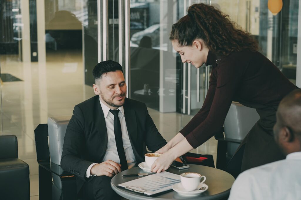 Optimistic bearded Caucasian businessman in formal clothes smiling and discussing business profit with his African American colleague in cafe at lunch time. Cheery waitress bringing cup of coffee.
